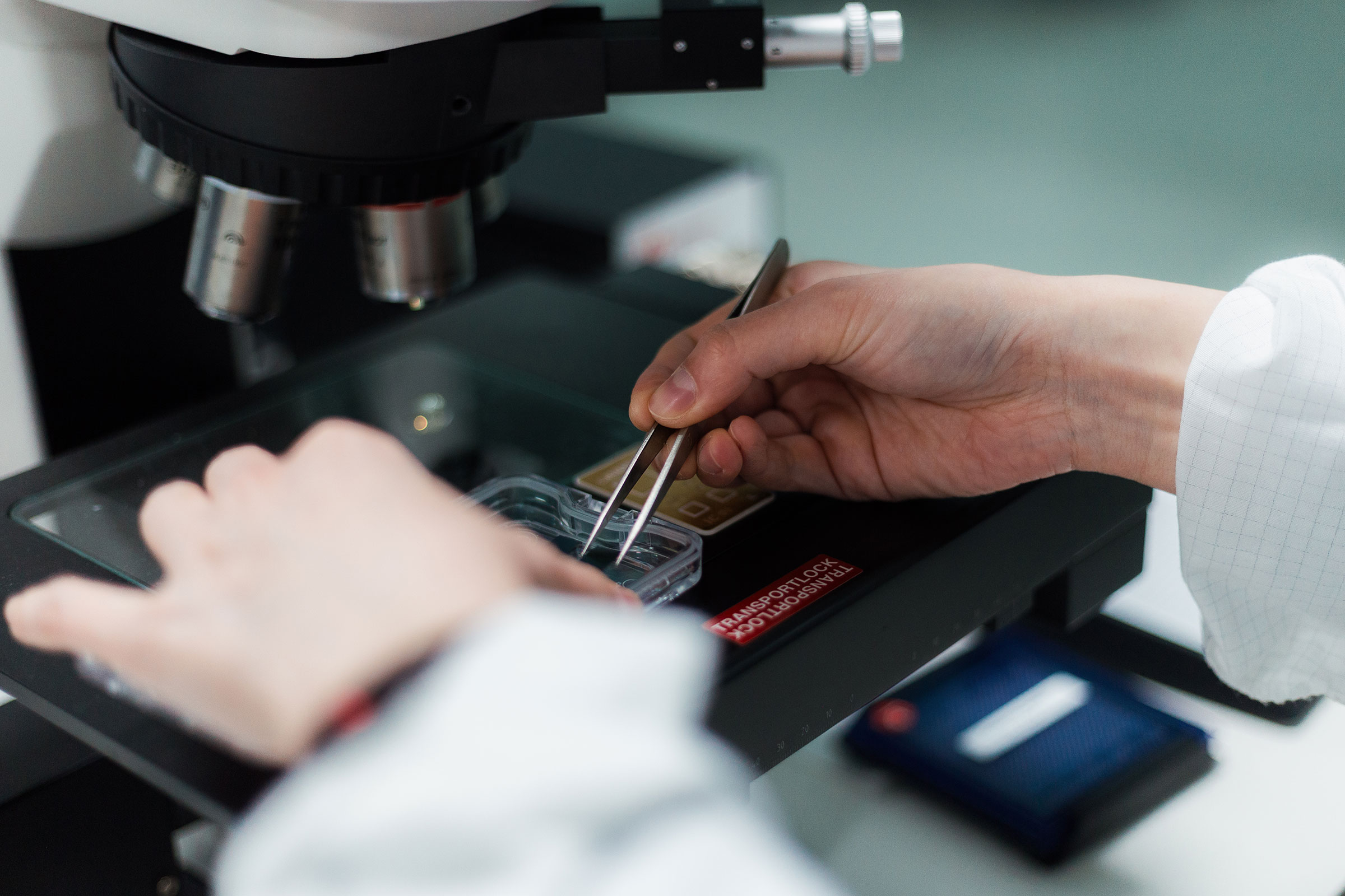 Close-up of hands using tweezers to handle a semiconductor chip under a microscope in a laboratory setting. - CSA Catapult
