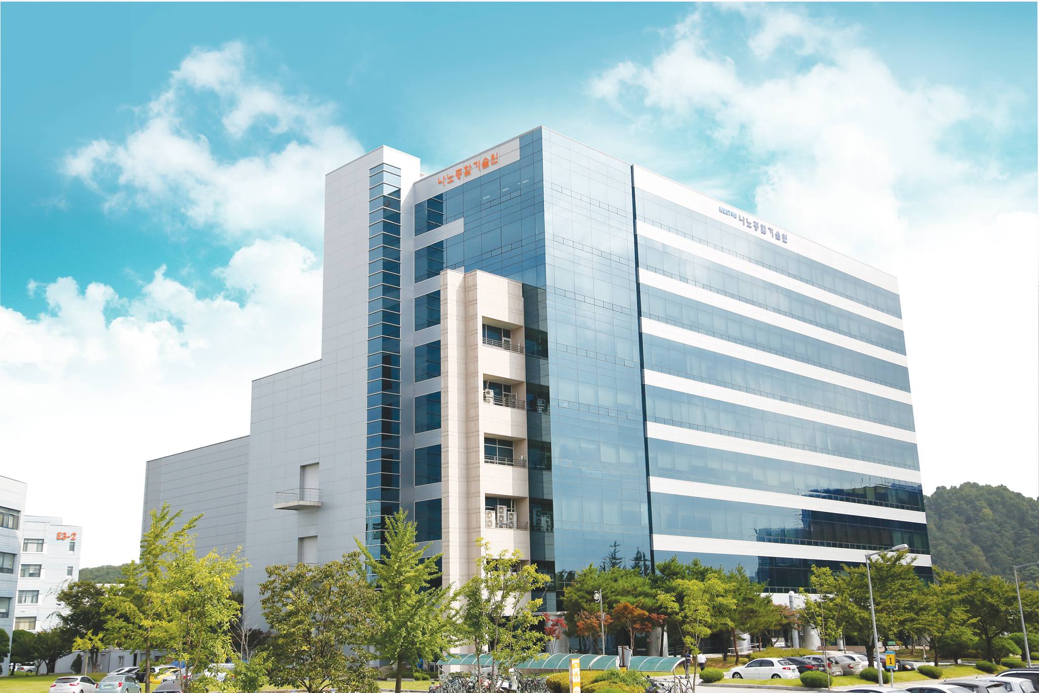 Modern glass office building, the National Nanofab Centre in Daejon, with Korean signage, trees, cars, and a blue sky. - CSA Catapult