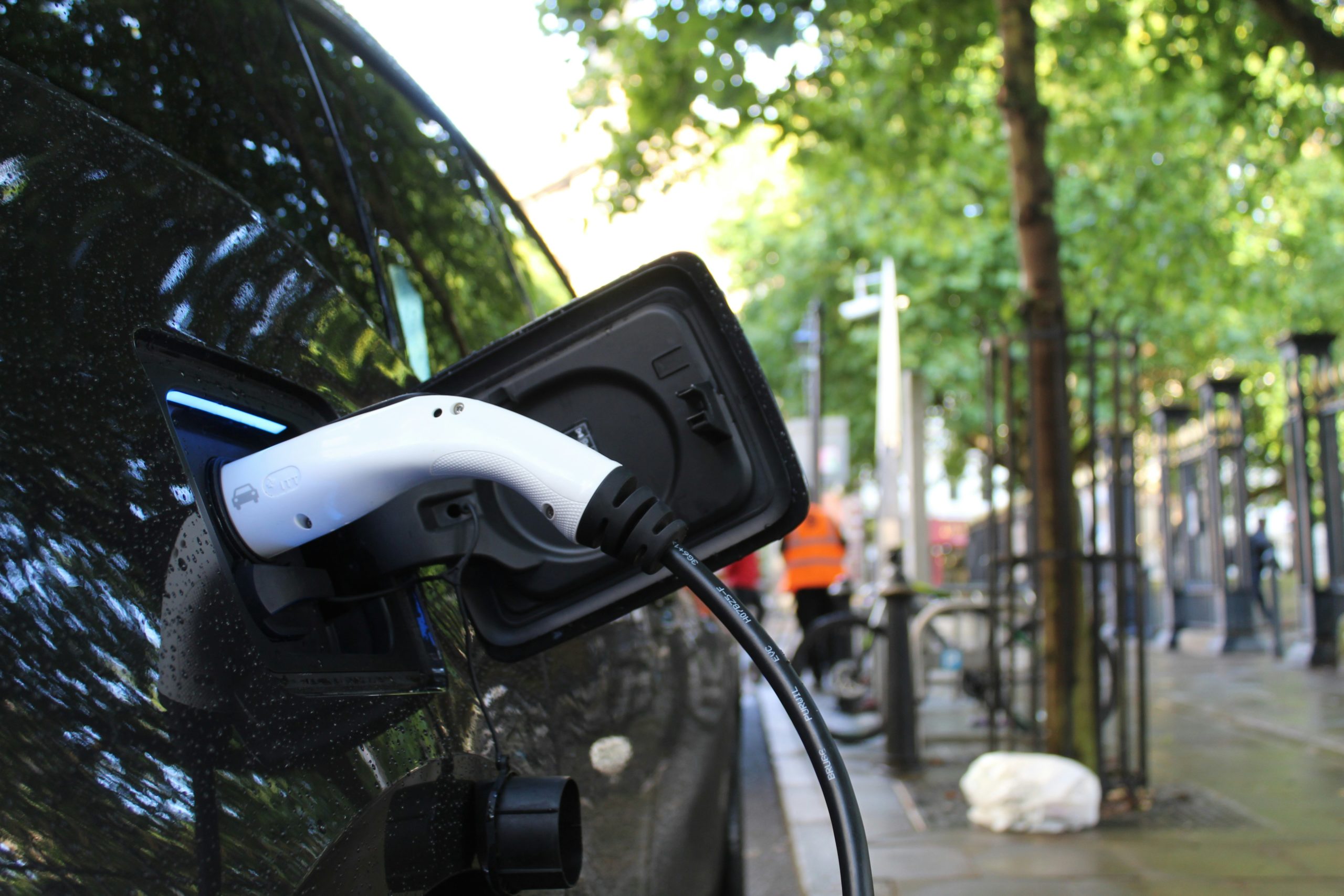 Close-up of an electric car charging on a city street, with AI-powered systems, trees, and a bike in the background. - CSA Catapult