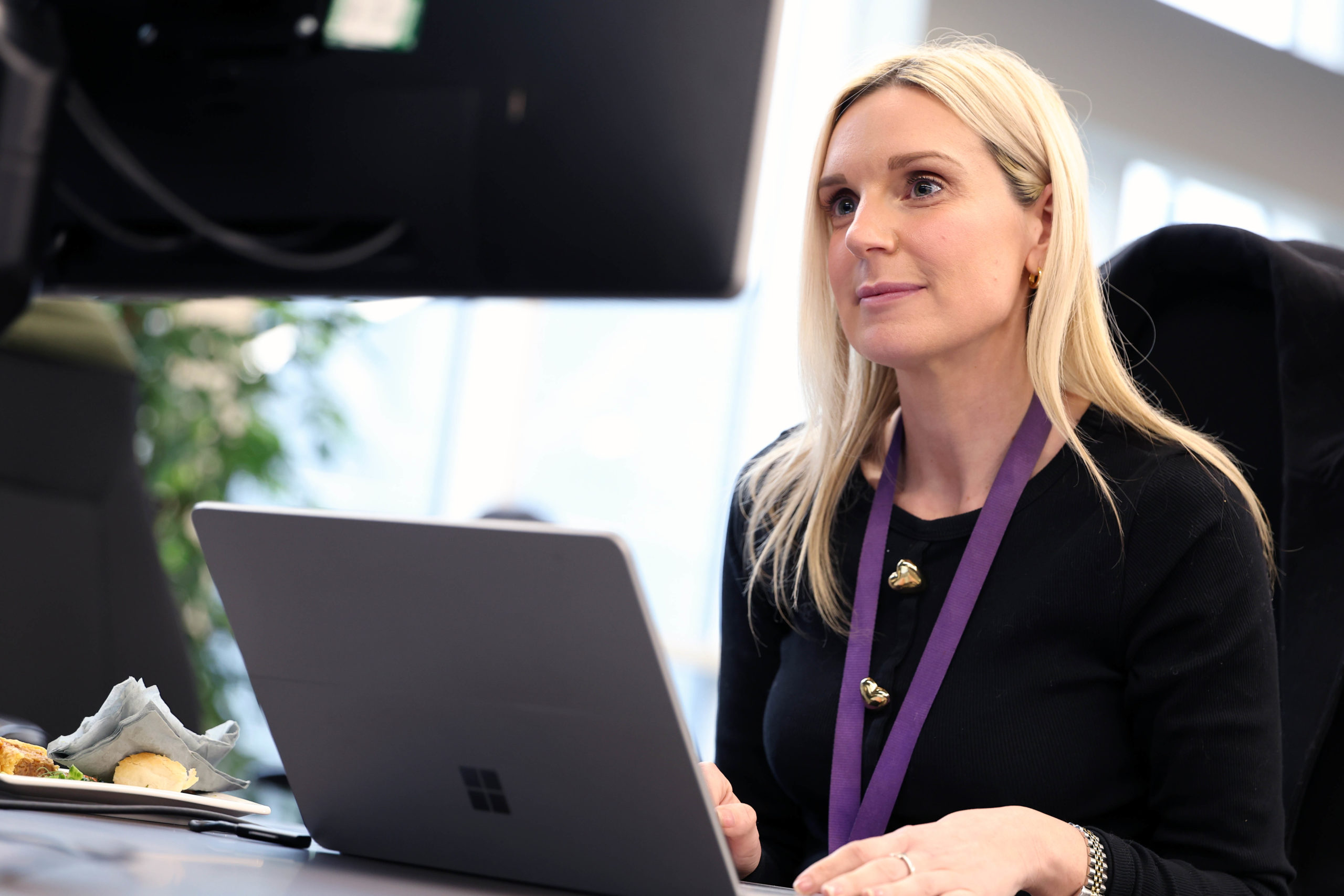Woman with long blonde hair working at a desk on a laptop, looking at a monitor, wearing a purple lanyard. - CSA Catapult