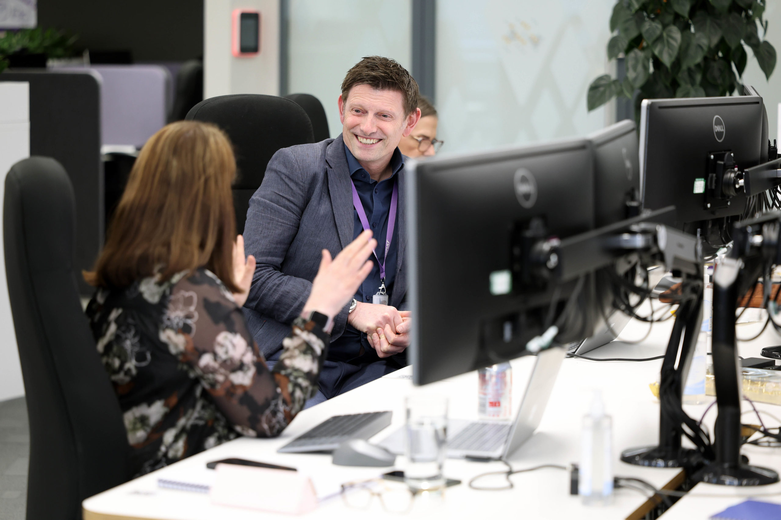 Two colleagues sitting at desks in an office, smiling and having a lively conversation by computer monitors. - CSA Catapult