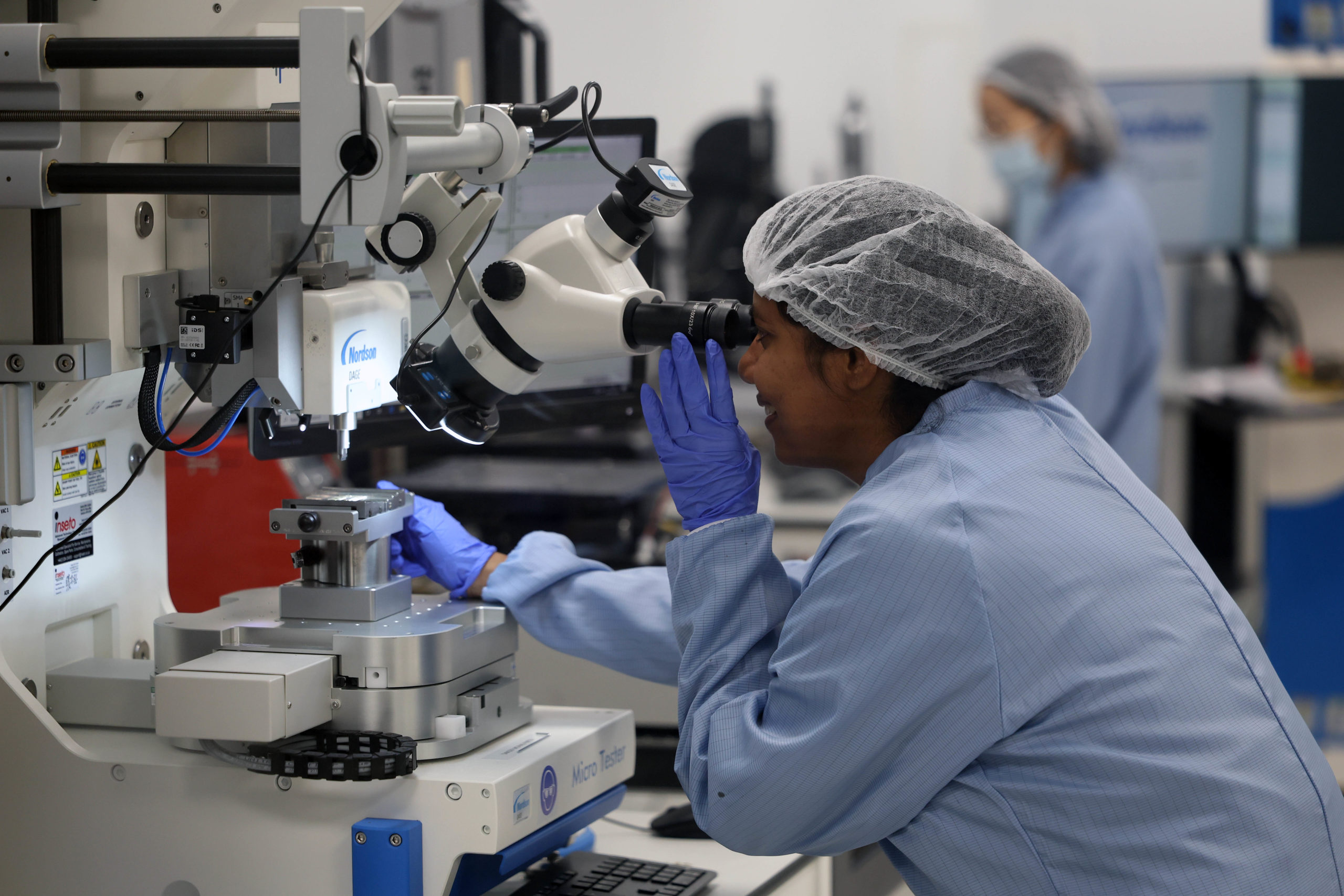 A scientist in a lab coat and hair net uses a microscope in a laboratory setting. - CSA Catapult