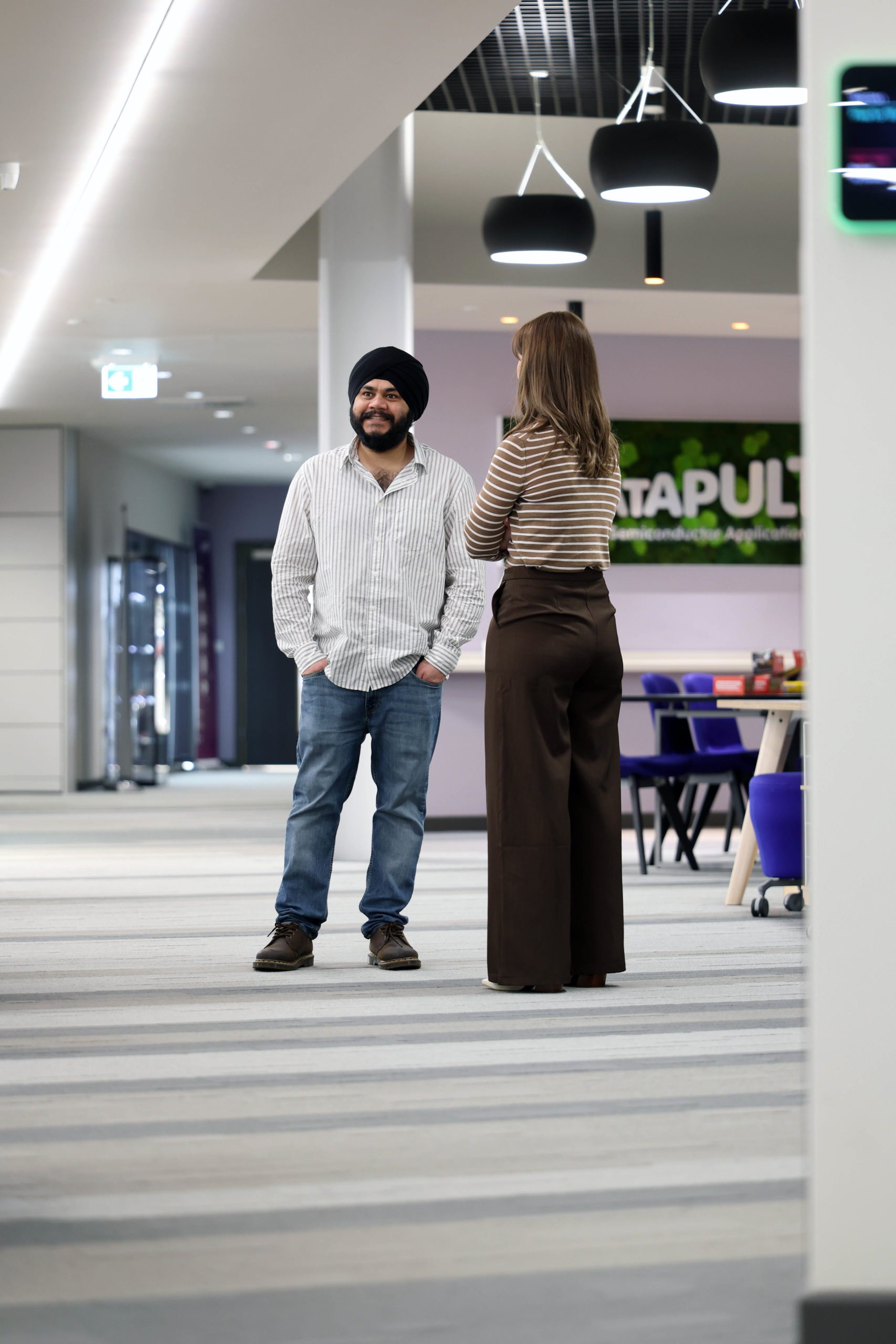 A man and a woman stand talking in a modern office hallway with desks and lights in the background. - CSA Catapult
