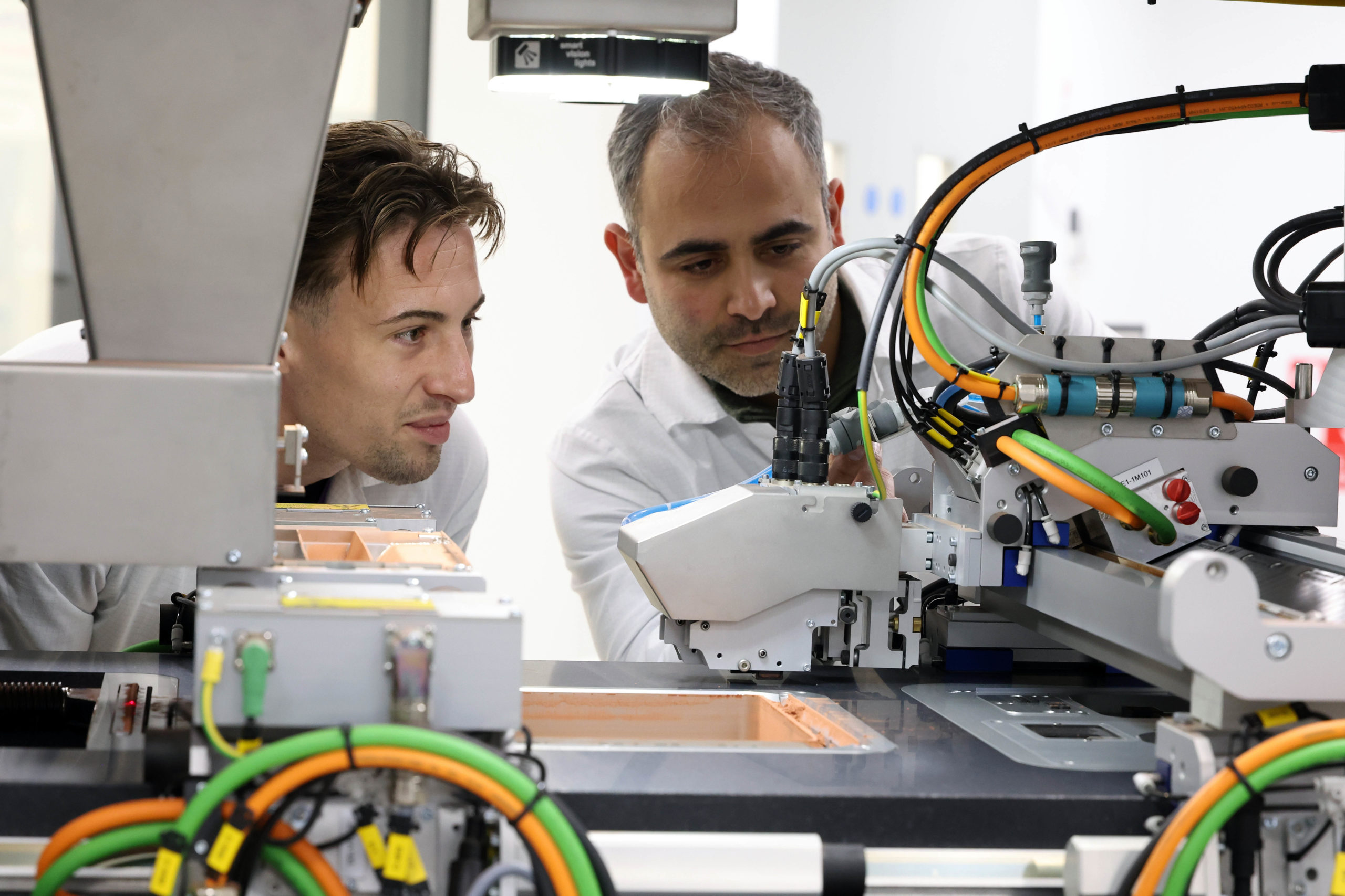 Two men closely observe and work on a machine with various wires and components in a lab setting. - CSA Catapult