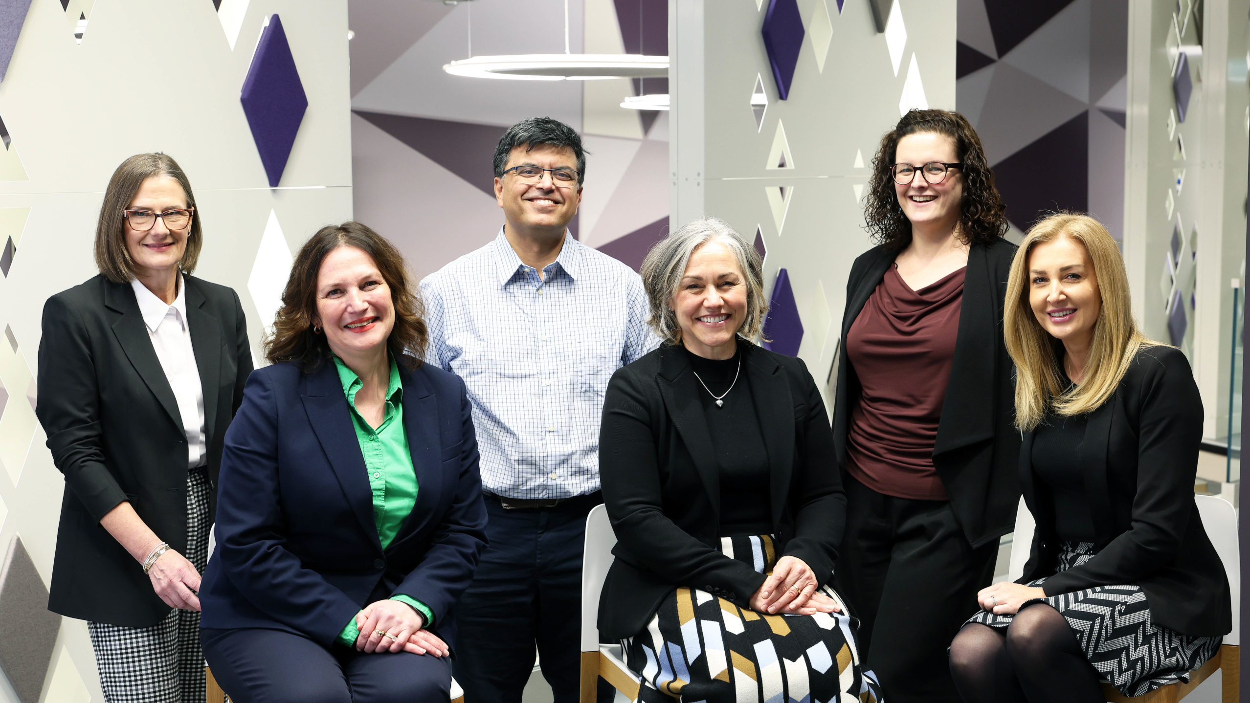 Six professionally dressed people pose together and smile in a modern office with geometric wall patterns. - CSA Catapult