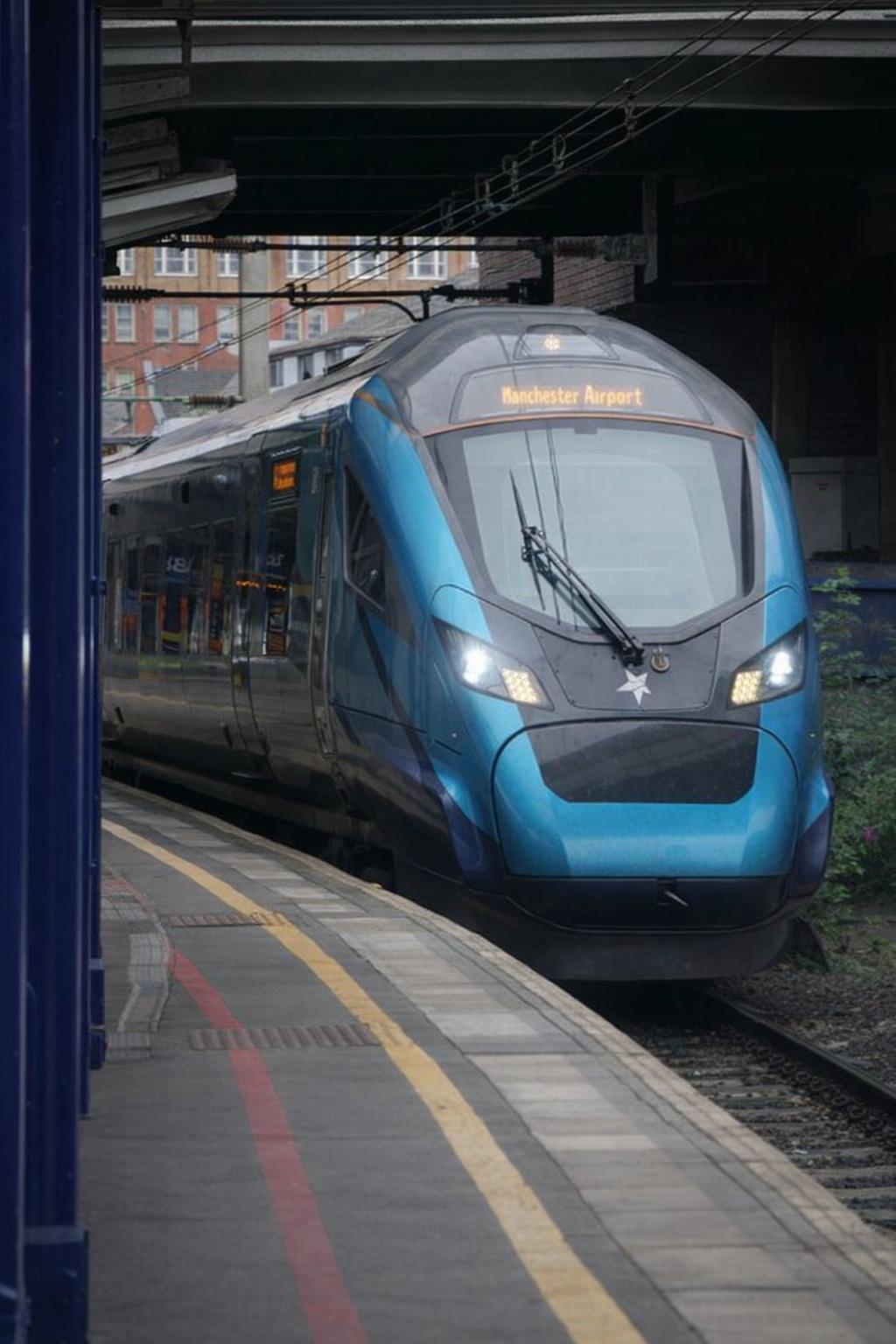 An AI-powered blue train to Manchester Airport approaches a platform under a bridge at a city railway station. - CSA Catapult