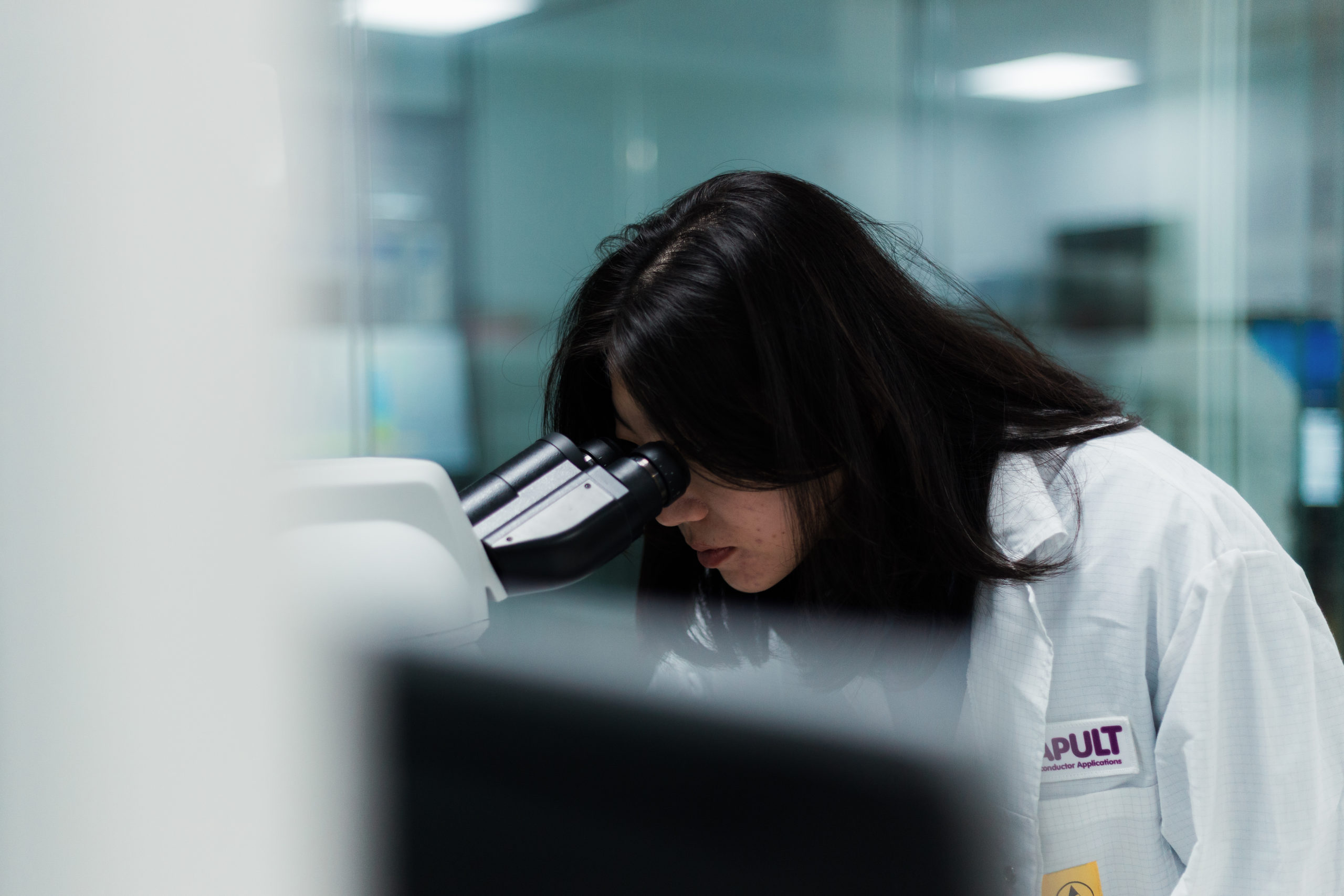 A person in a lab coat looks through a microscope in a laboratory setting. - CSA Catapult
